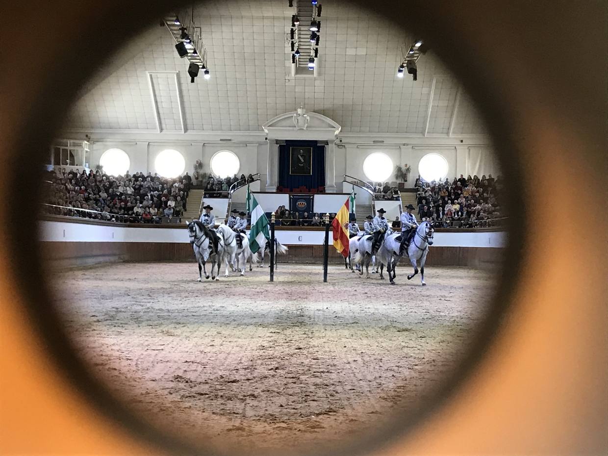 Le carrousel de la Real Escuela vu par un judas : des cavaliers en costume andalou sur des chevaux gris, entre les drapeaux andalou et espagnol, devant un public nombreux.